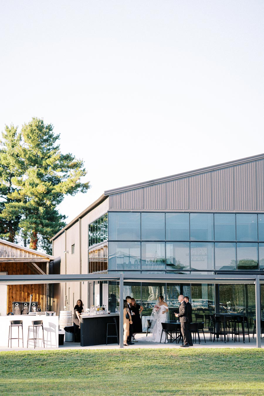 Exterior view of a modern glass event venue with guests mingling and a bar setup, surrounded by lush greenery and large trees on a sunny day.