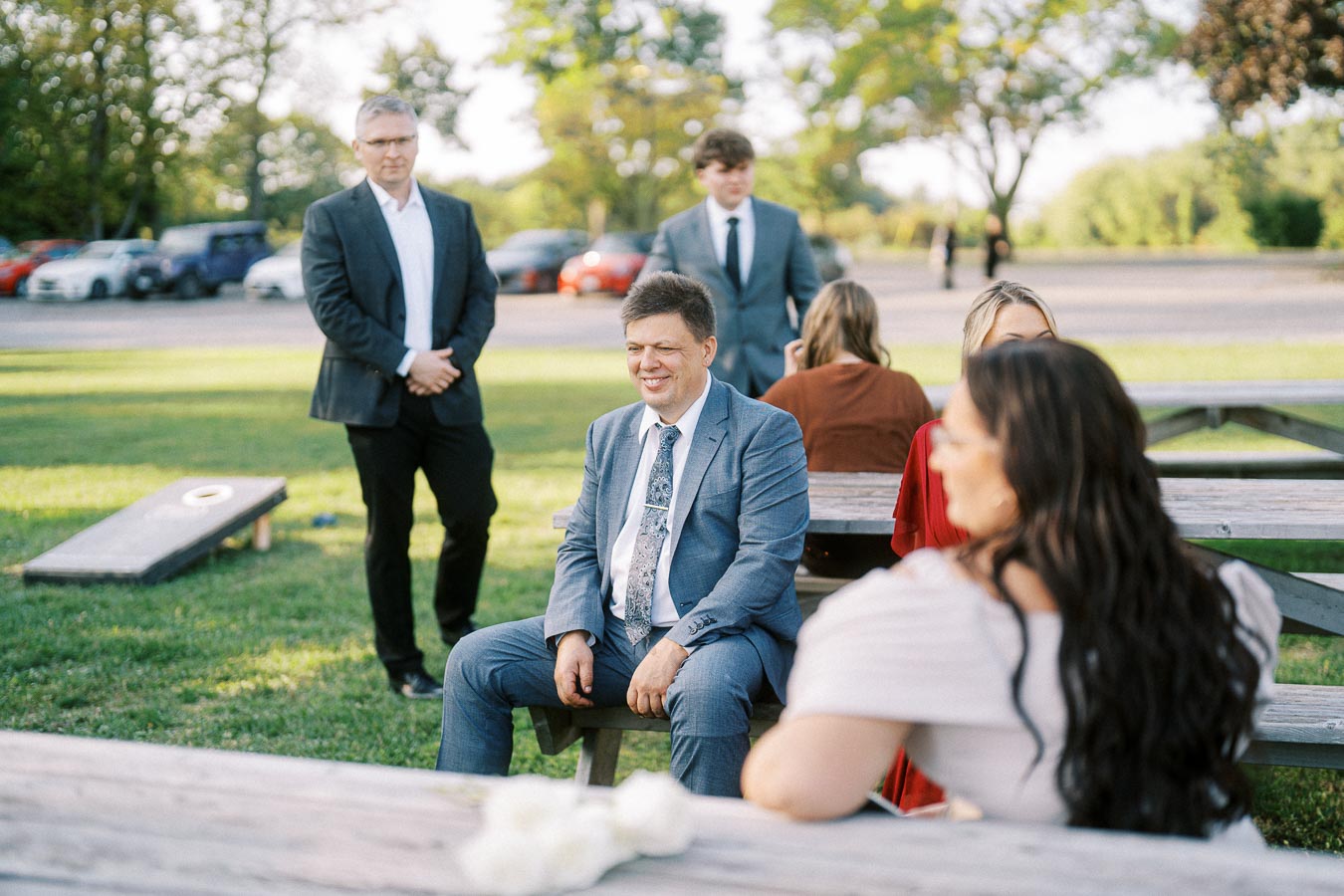 A group of people in formal attire socializing outdoors, seated at picnic tables on a sunny day, with green grass and trees in the background.