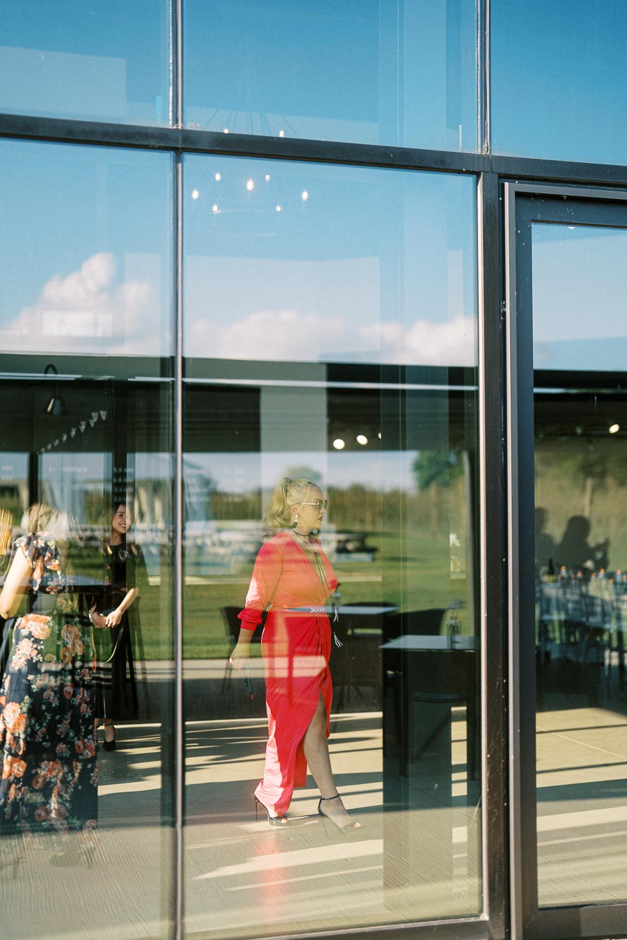 Stylish woman in a red dress walking indoors, reflected in a glass window, with outdoor scenery visible in the background.