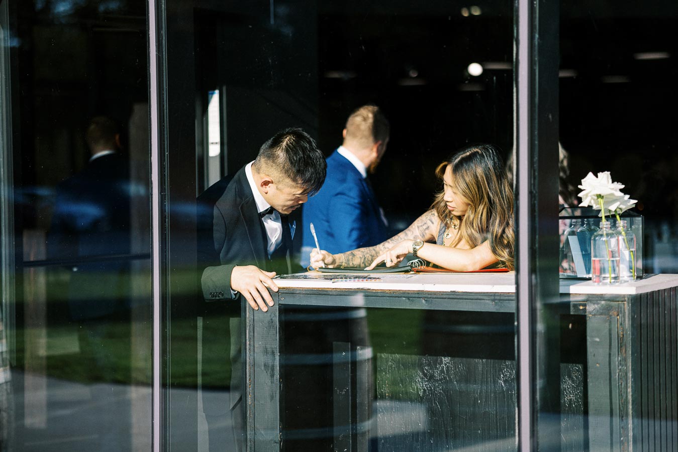 A man and woman in formal attire signing a guest book at an elegant event venue, with a vase of white flowers on the table.