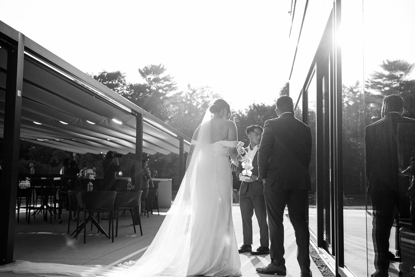 Black and white image of a bride in a flowing wedding dress and veil standing with two men in suits outside a modern building during a sunny day, capturing a candid wedding moment.