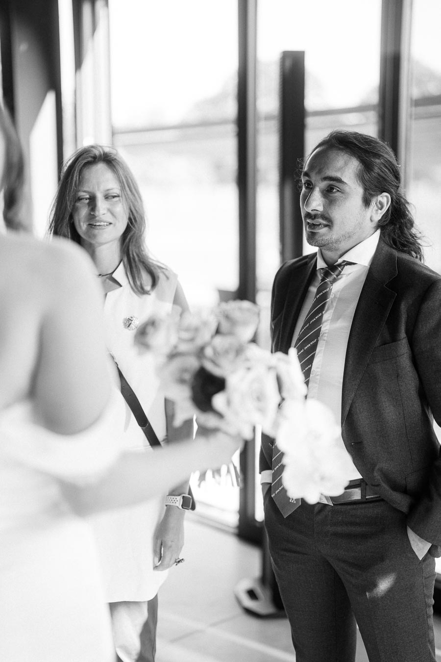 Black and white image of two people conversing with a bride holding a bouquet, set in a brightly lit room with large windows.