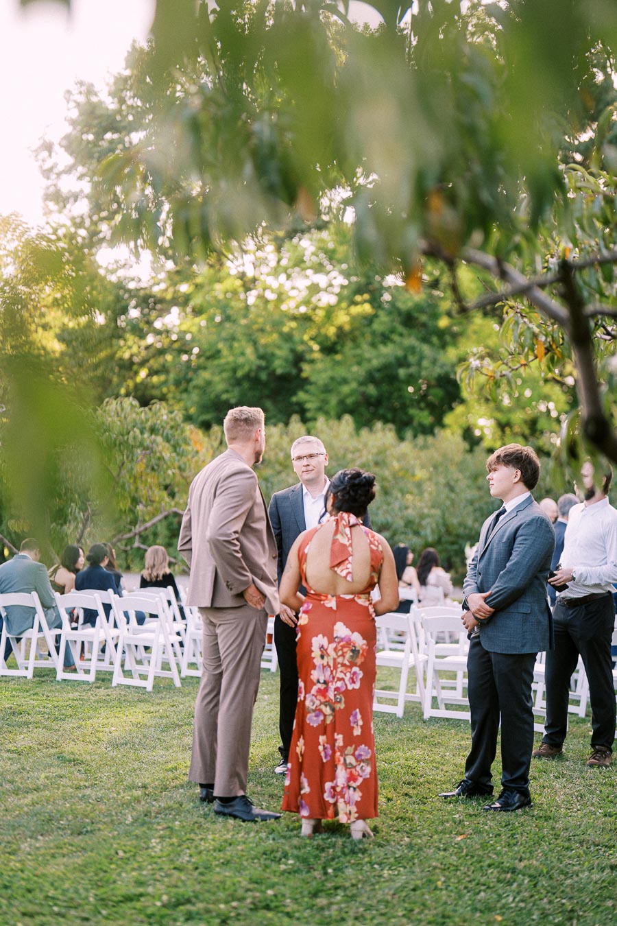 A group of well-dressed individuals engaged in conversation at an outdoor wedding venue, surrounded by lush greenery and white folding chairs, capturing a candid moment amidst a garden setting.