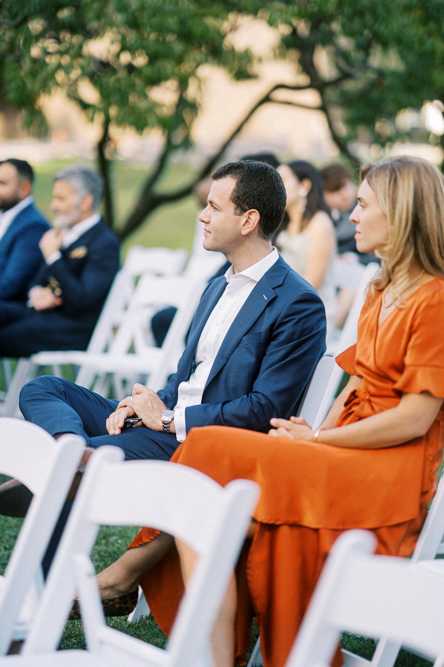 Guests seated outdoors at a formal event, with a man in a blue suit and a woman in an orange dress, attentively listening, surrounded by white chairs and greenery.