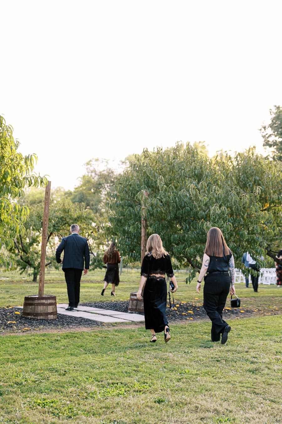 People in formal attire walking through a lush garden pathway during a gathering or event, surrounded by green trees and grass under a clear sky.