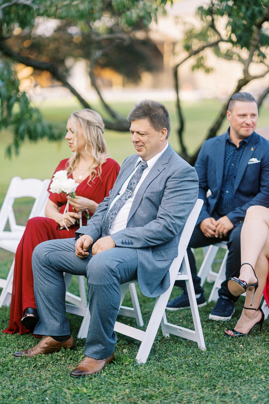 Outdoor wedding guests seated in white chairs, including a man in a gray suit and a woman in a red dress holding white flowers, amidst a green garden setting.