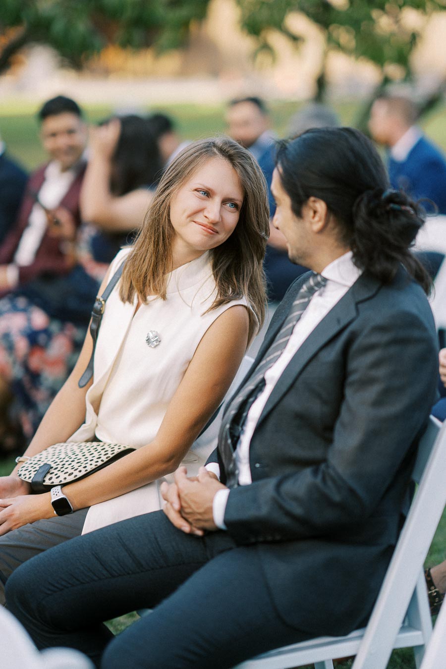 A woman in a white sleeveless top smiling at a man in a suit and tie at an outdoor event. They are sitting on white chairs with other attendees visible in the background. The scene is set in a garden with greenery and blurred party guests, providing an elegant atmosphere.
