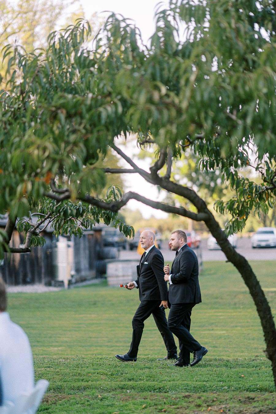 Two men in suits walking on a grassy lawn under a tree at an outdoor event, with parked cars and a rustic building visible in the background.