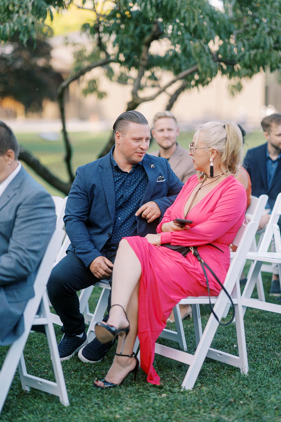 A man in a blue suit sits conversing with a woman in a vibrant pink dress at an outdoor event. They are seated on white folding chairs on a grassy lawn, with other attendees visible in the background.