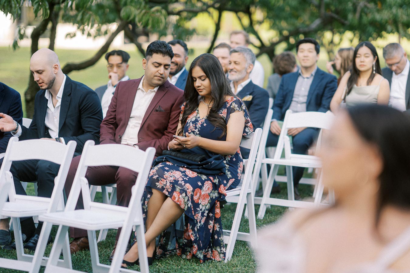 A diverse group of people seated in white chairs at an outdoor gathering, some engaged in conversation while others focus on their phones, surrounded by greenery and trees.