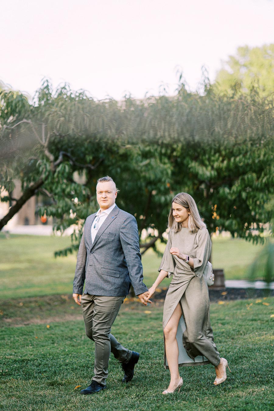 A couple walking hand in hand through a lush garden setting, the man wearing a gray suit and the woman in an elegant olive dress, capturing a moment of elegance and connection.