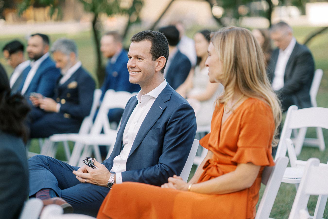 Attendees seated at an outdoor event, with a man in a blue suit and a woman in an orange dress, engaging in conversation and smiling.