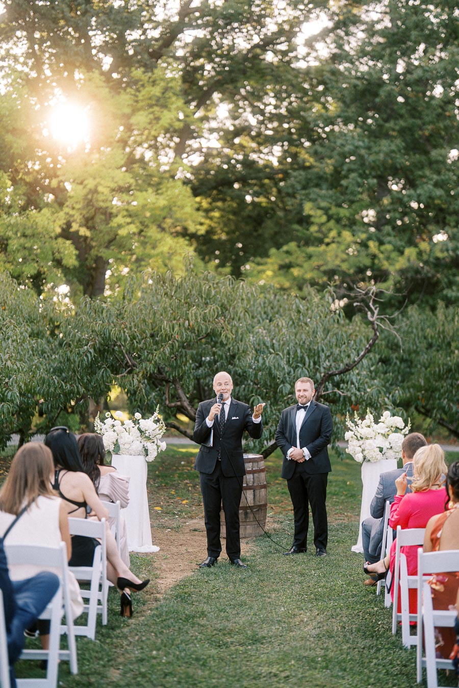Outdoor wedding ceremony with a man giving a speech in a lush garden setting, surrounded by guests seated in white chairs and floral decorations.