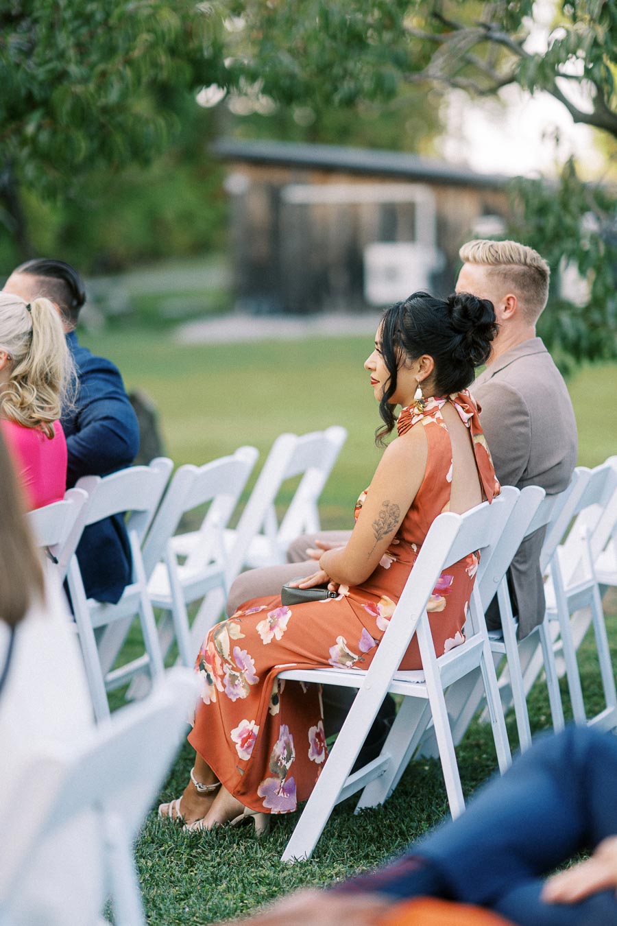 Guests seated on white chairs at an outdoor wedding ceremony, woman in floral dress listening attentively.