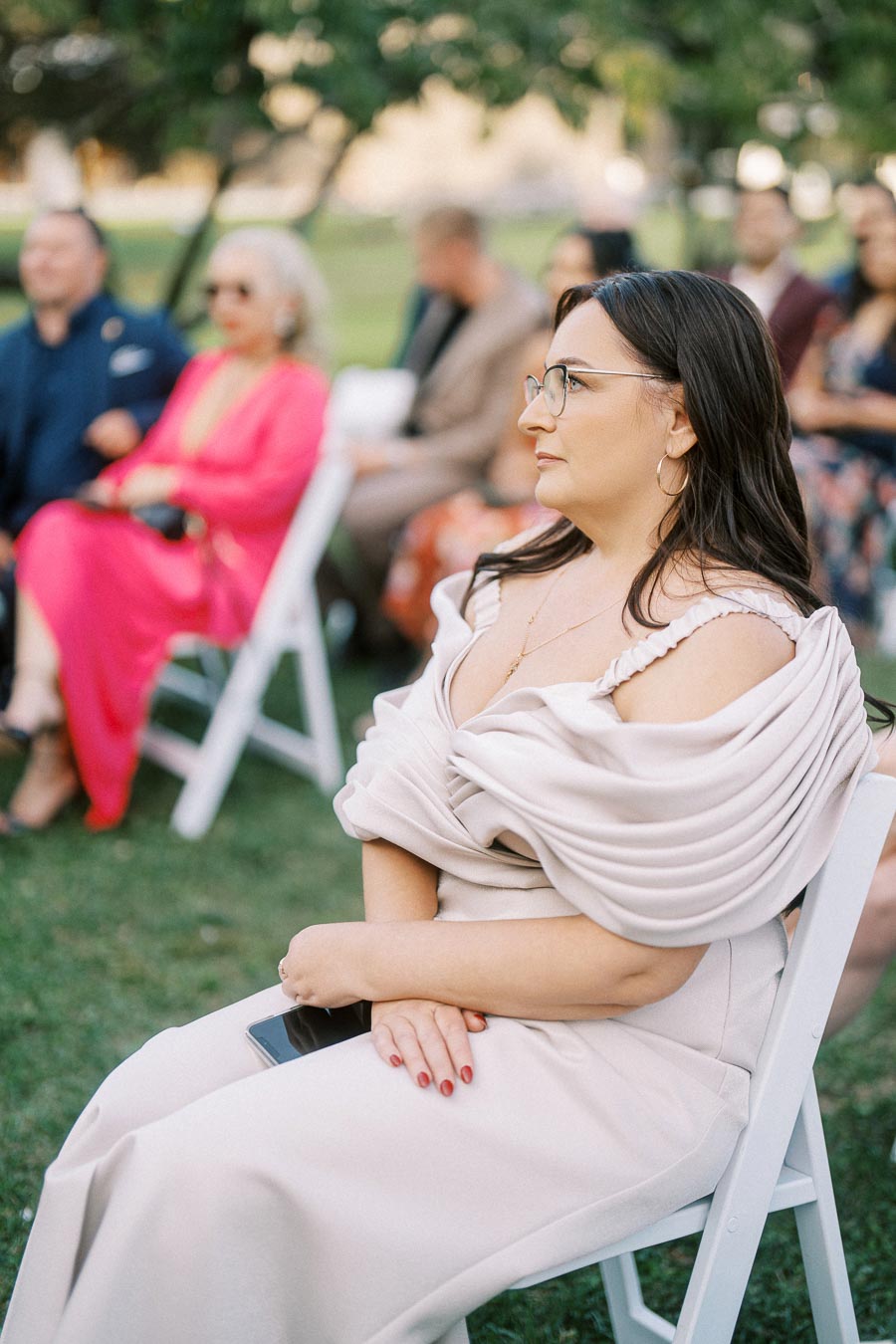 A woman in a stylish, off-the-shoulder dress with glasses sits attentively at an outdoor event with other attendees in the background, surrounded by green foliage.