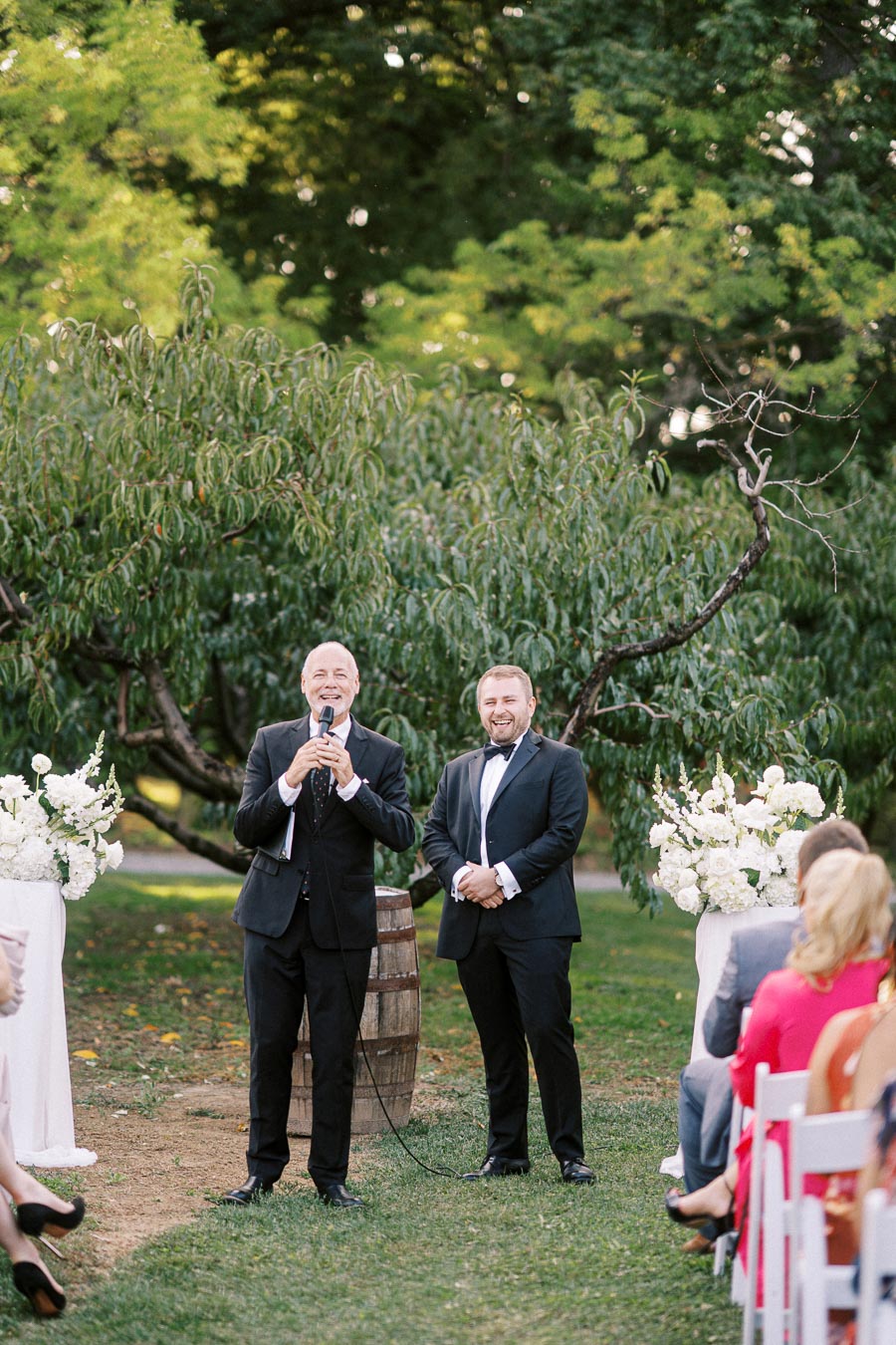 Outdoor wedding ceremony with two men in suits standing in front of guests. One man is speaking into a microphone while the other smiles. They are surrounded by lush greenery and decorative white floral arrangements.