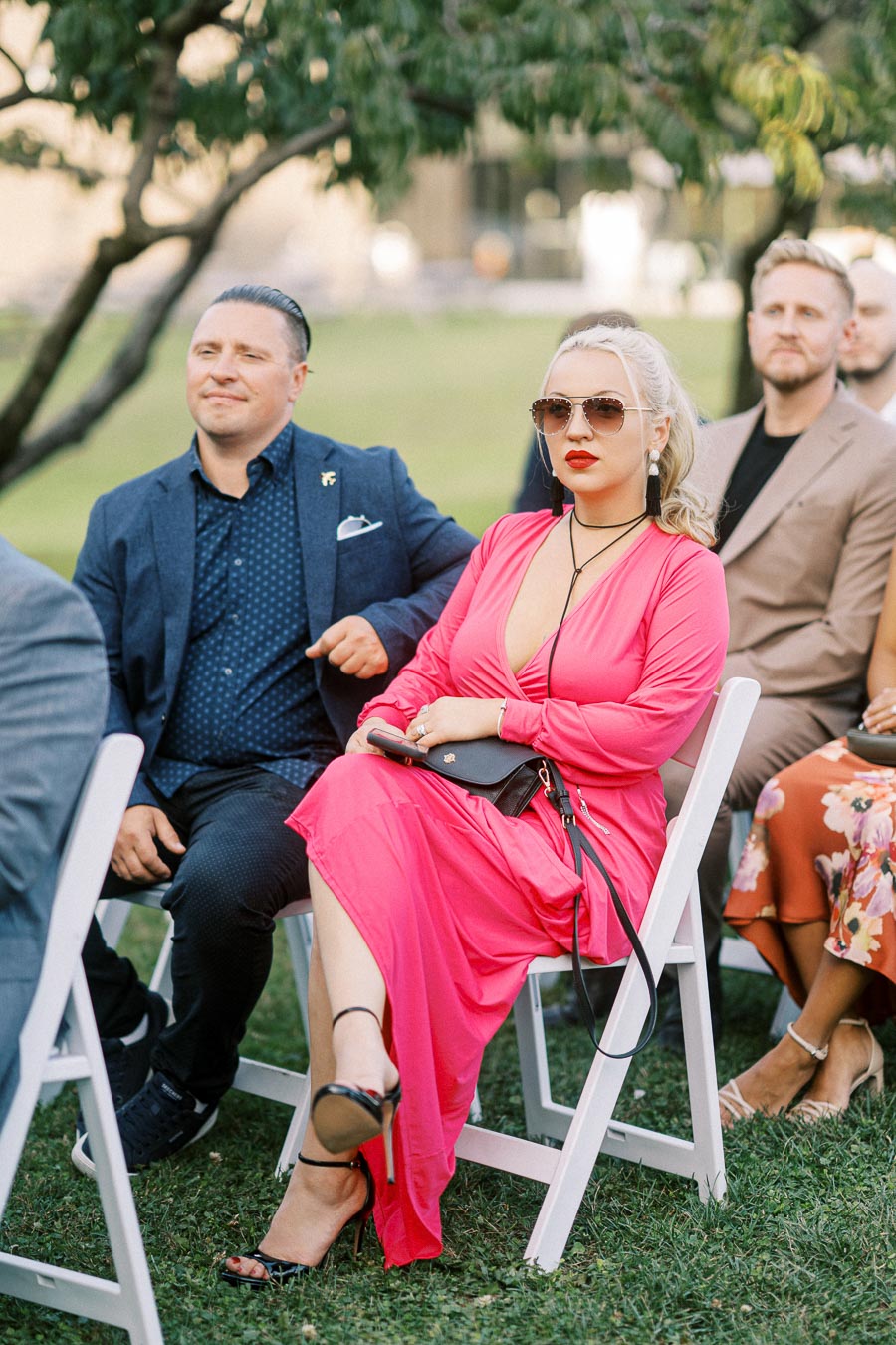 A stylish woman in a bright pink dress and sunglasses sitting on a white chair outdoors at a formal event, accompanied by a man in a navy suit, with other attendees in the background.
