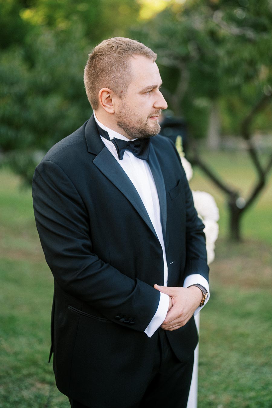 A man in a black tuxedo and bow tie standing outdoors on a grassy lawn, looking to the side with trees in the background.