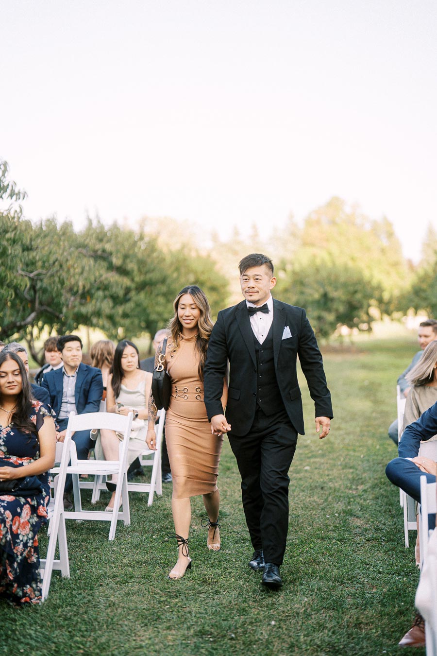 Elegant couple walking down outdoor aisle during wedding ceremony, surrounded by seated guests in a scenic garden setting