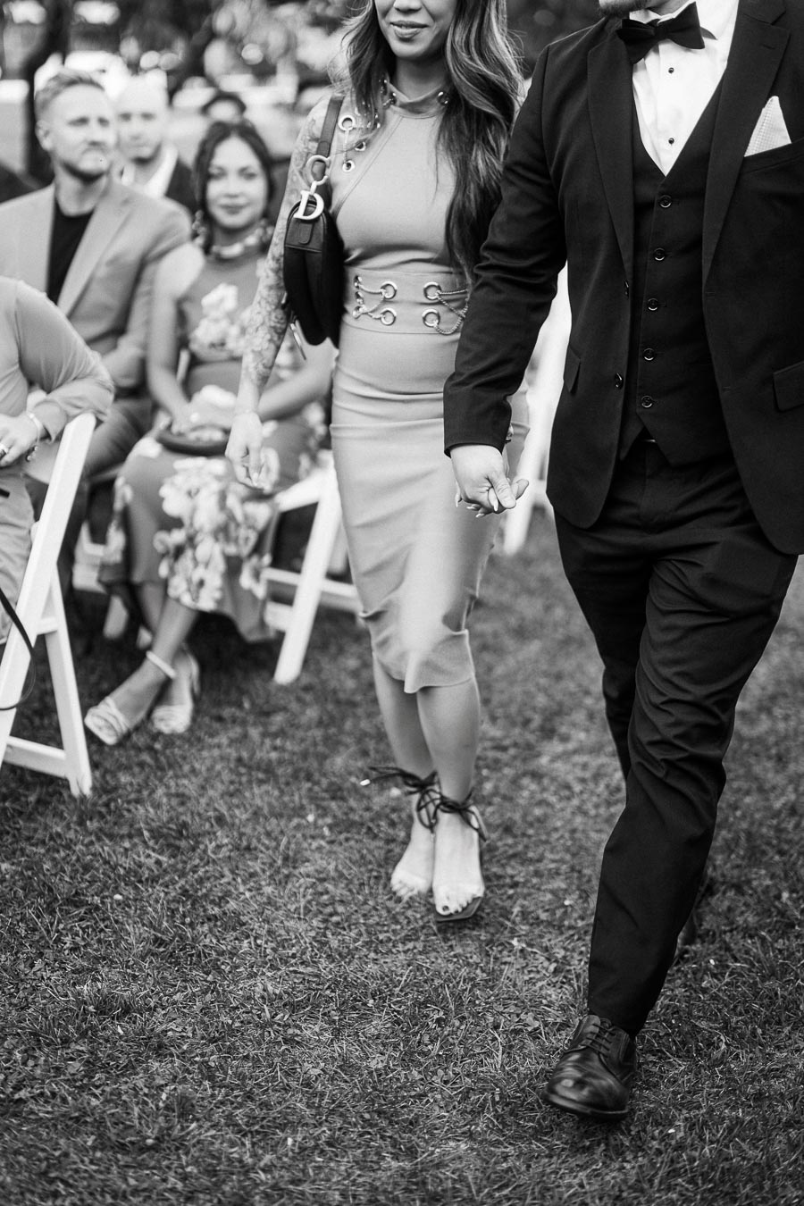 A black and white image depicting a stylishly dressed couple walking hand in hand down an aisle at an outdoor event, with seated guests in the background, showcasing elegant fashion and a sense of occasion.