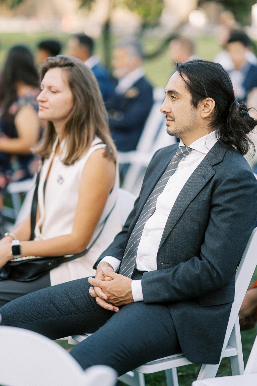Two people sitting on white chairs outdoors at an event, wearing business attire.