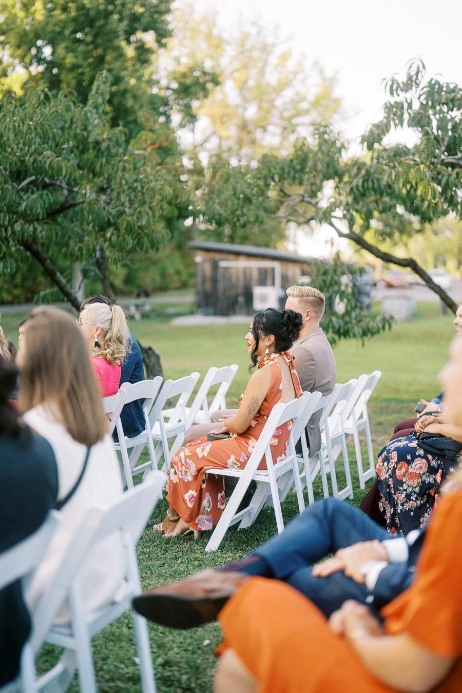 Outdoor wedding ceremony with guests seated on white chairs, surrounded by lush trees and greenery.