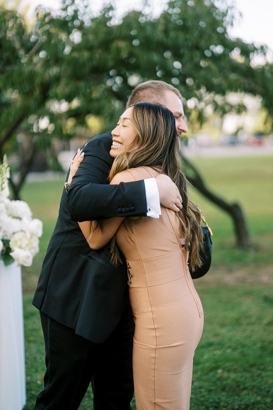 A woman in a beige dress hugging a man in a black suit in a garden setting, with trees and flowers in the background.