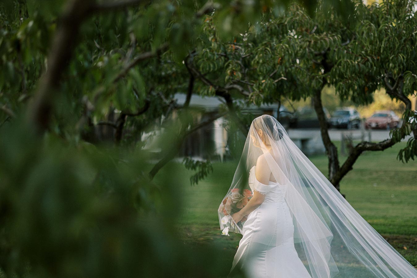 A bride in a white gown and veil holding a bouquet walks through a lush garden, surrounded by greenery and trees.