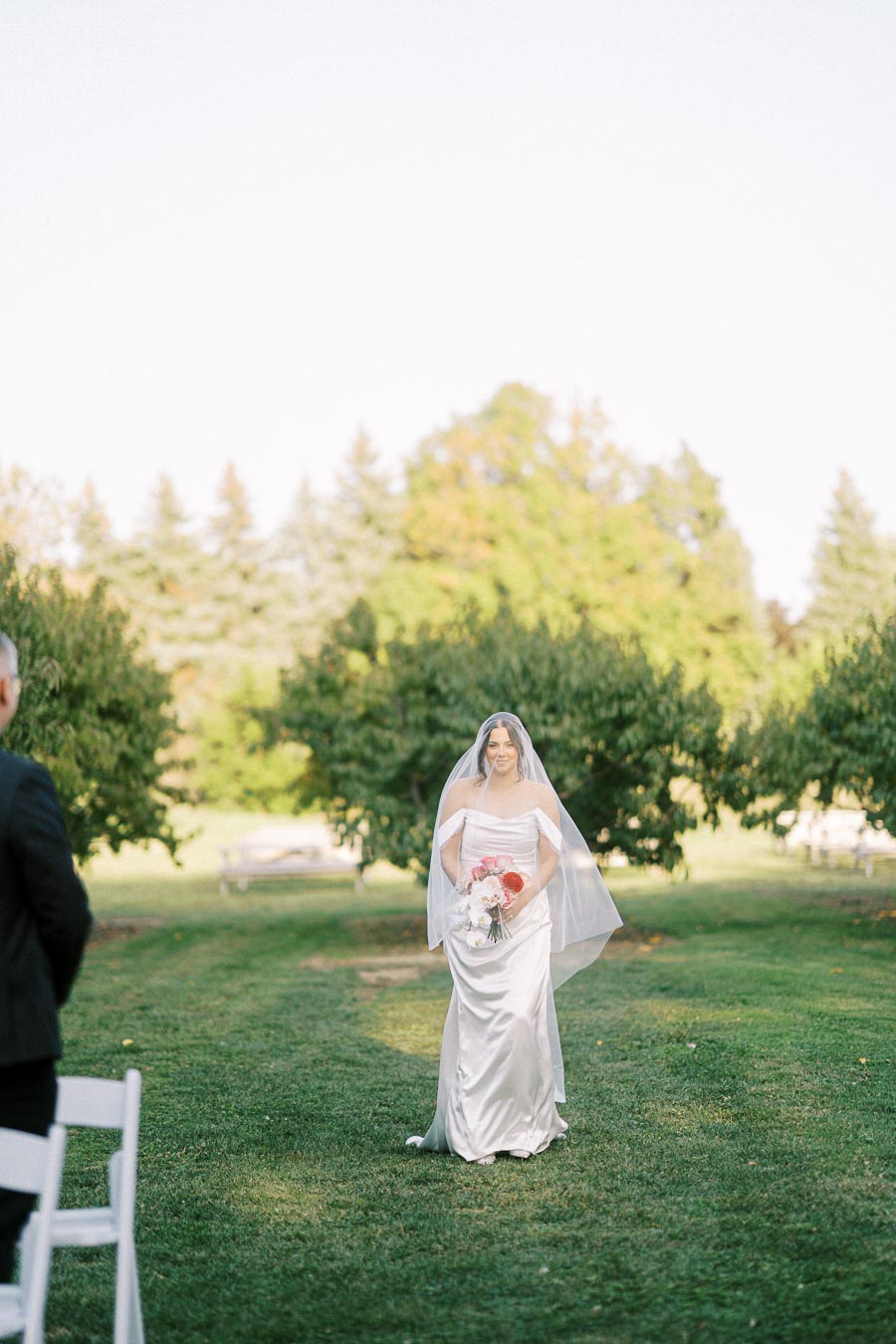 Bride in white wedding dress and veil holding a bouquet, walking through a scenic outdoor garden setting with green trees and grass.