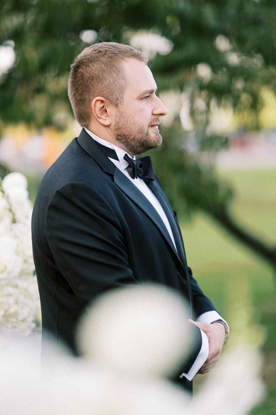 Man in a tuxedo standing outdoors with a blurred green background, looking thoughtful.