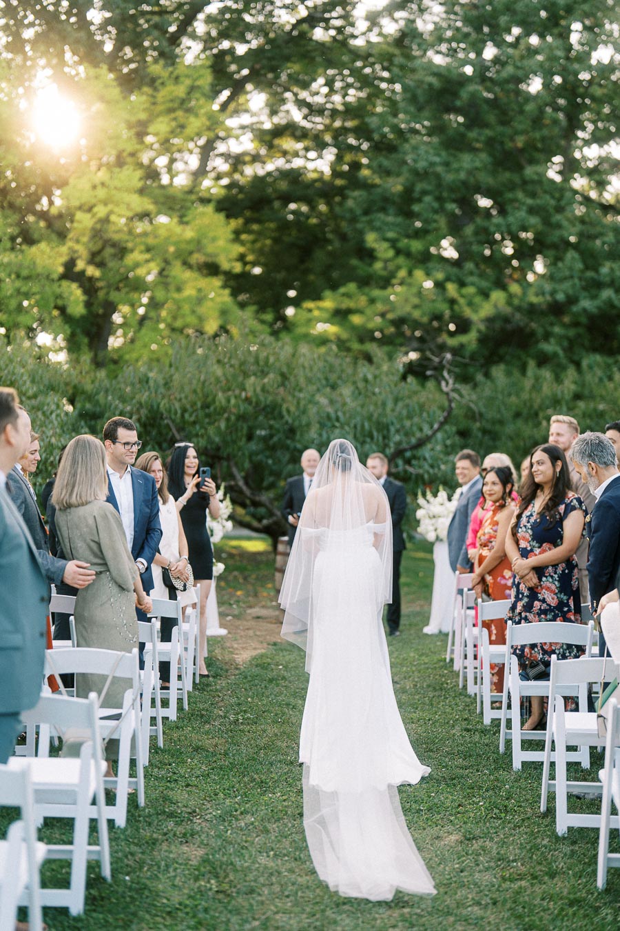 Bride walking down the aisle at an outdoor wedding ceremony surrounded by guests on a sunny day.