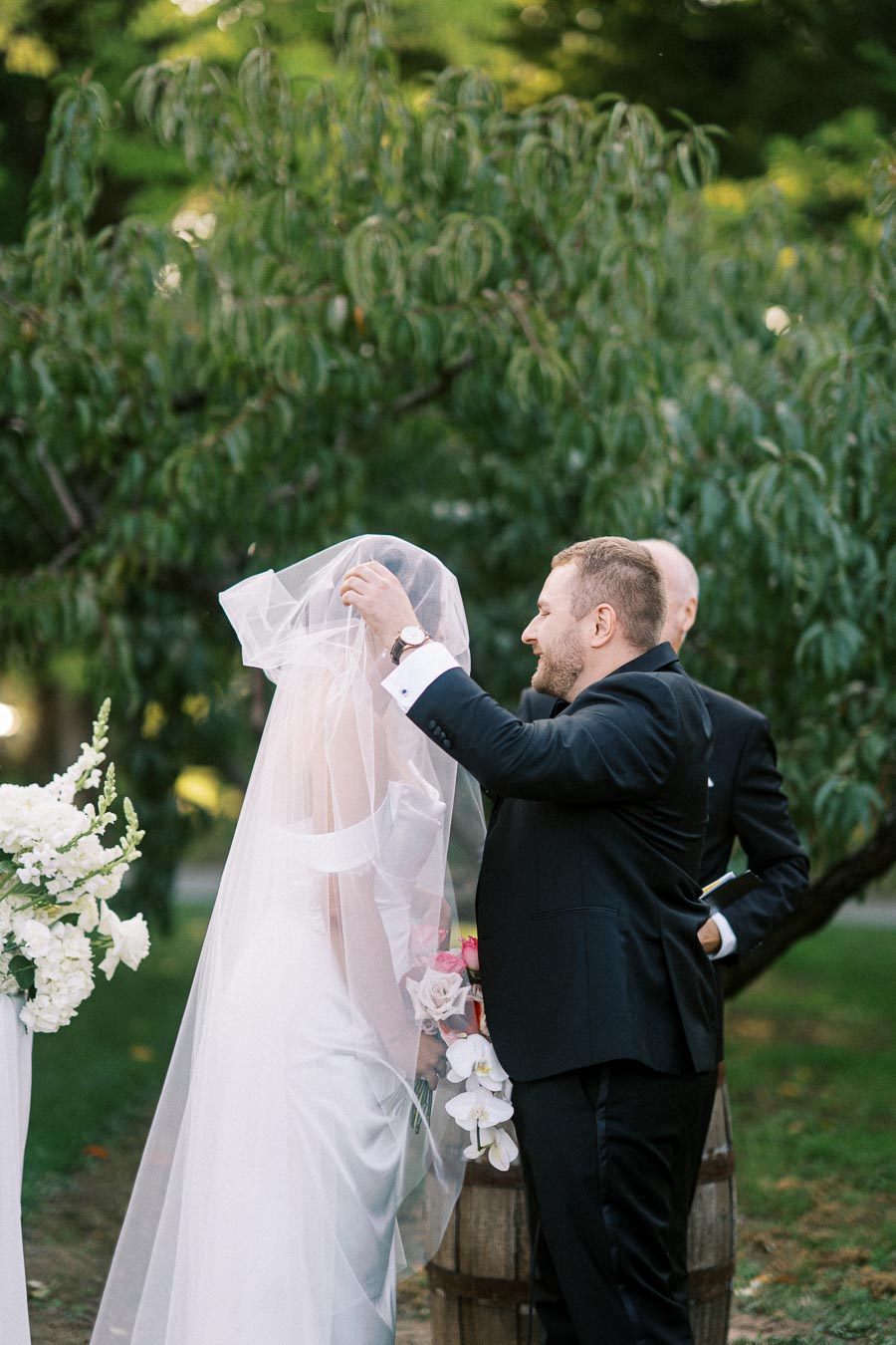 A groom lifts the bride's veil during an outdoor wedding ceremony surrounded by lush green trees and white floral arrangements.