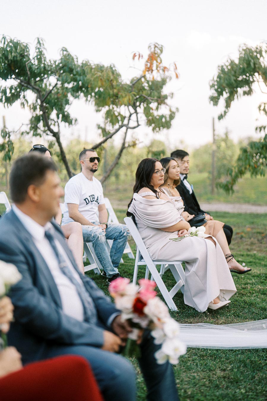 A group of elegantly dressed guests seated outdoors, surrounded by greenery and trees, attending a wedding ceremony on a sunny day.