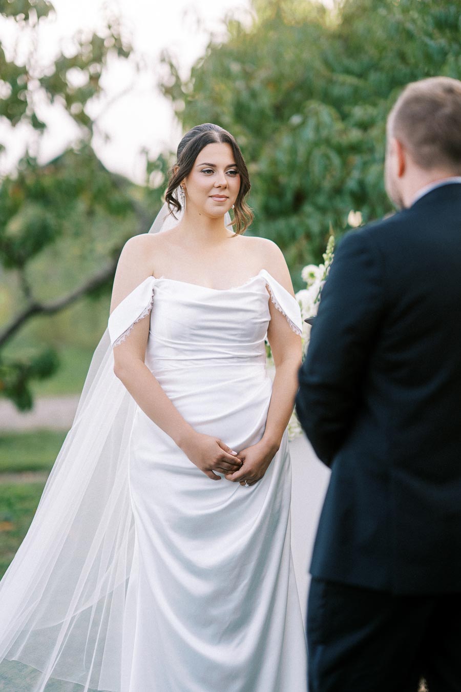 A bride in an elegant white wedding gown stands outdoors during a wedding ceremony, with trees in the background, facing a person in a suit.