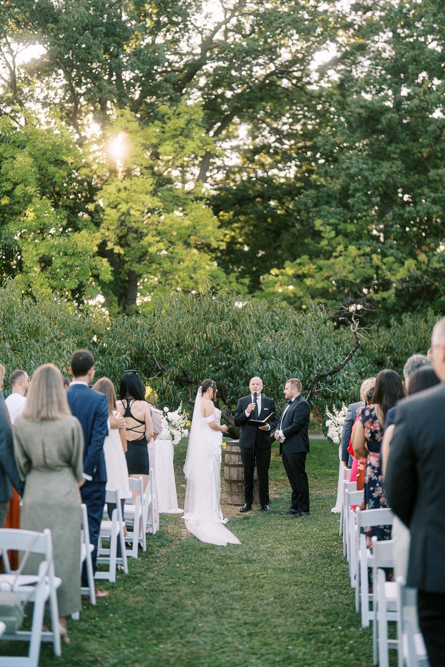 Outdoor wedding ceremony set in a lush garden with a couple exchanging vows under tall trees, surrounded by seated guests in a natural setting.