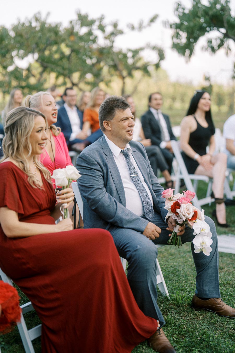 Attendees sitting outdoors holding flowers at a daytime wedding ceremony, dressed in formal attire with women in dresses and men in suits.