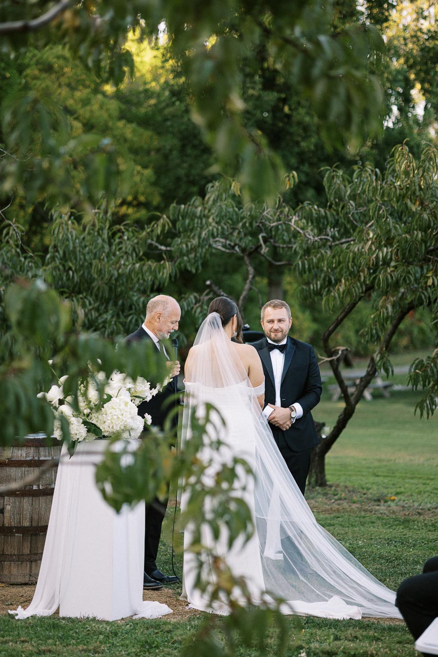 Wedding ceremony in a lush outdoor setting, with a couple exchanging vows under trees. The bride in a white gown with veil, and groom in a black tuxedo, stand in front of the officiant next to a floral arrangement.
