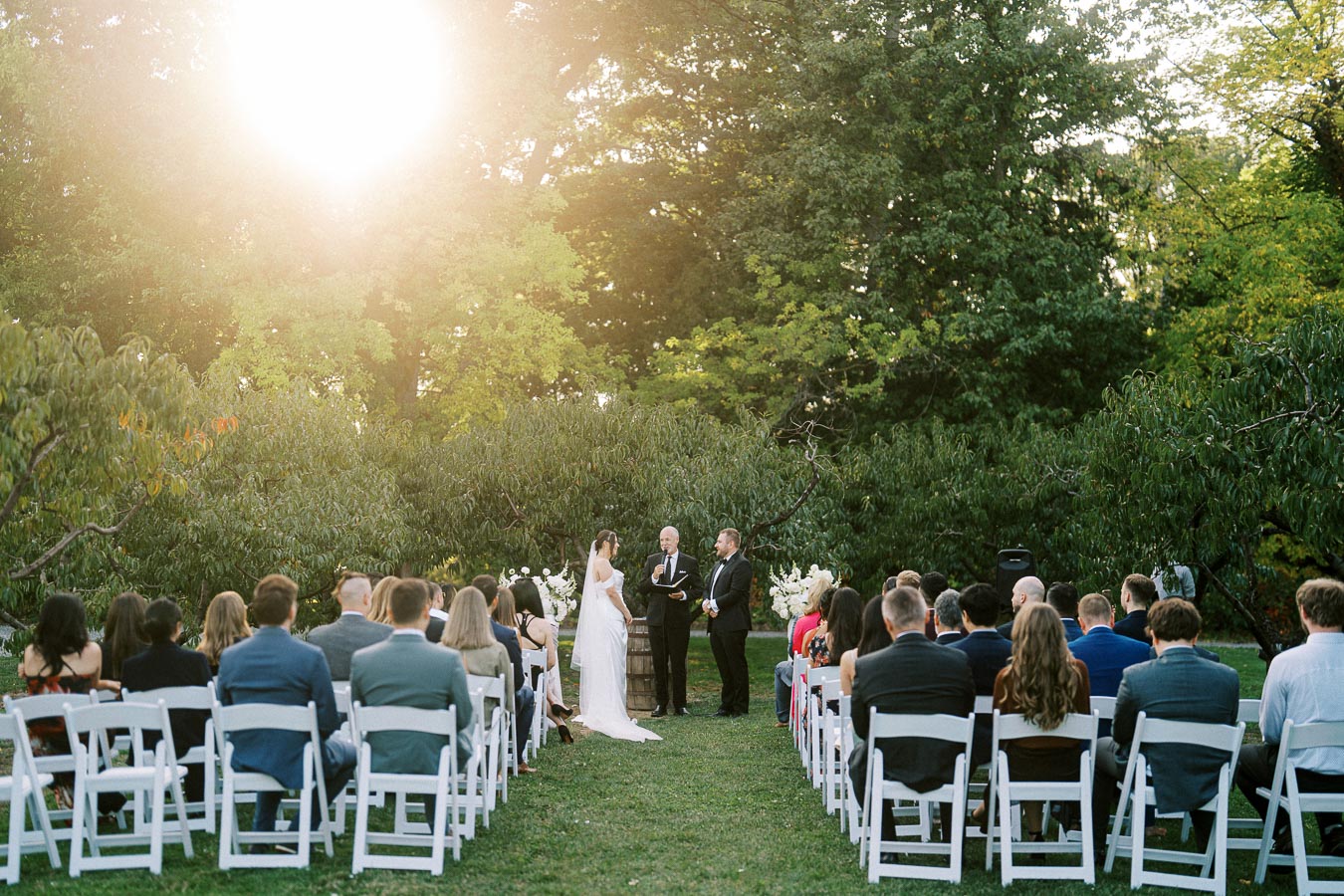 Outdoor wedding ceremony with a couple exchanging vows under the sunlight, surrounded by guests seated on white chairs in a lush garden setting.