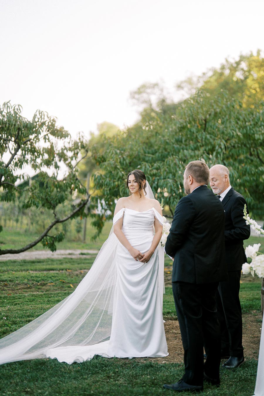 Bride in elegant white gown stands outdoors during wedding ceremony with officiant, surrounded by lush greenery.