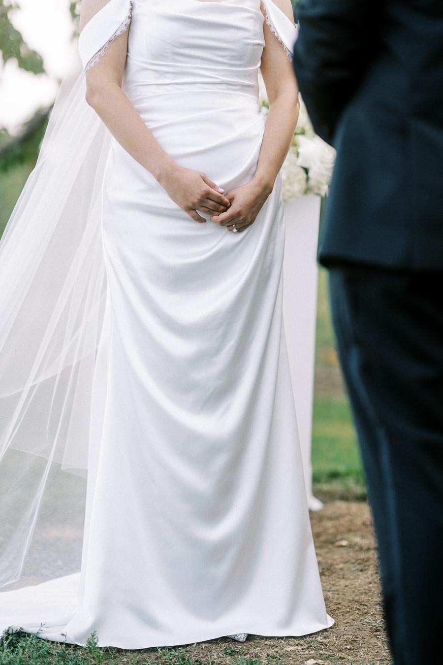 Bride in an elegant white wedding dress holding hands, standing outdoors during a ceremony, with a groom in a dark suit partially visible.