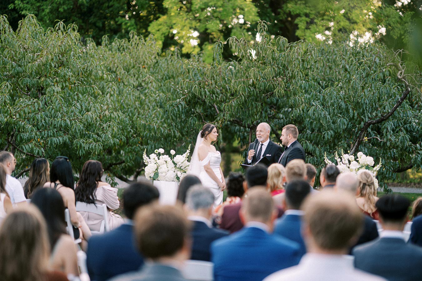 Outdoor wedding ceremony with a bride and groom standing in front of a floral arrangement, officiant speaking, and guests seated, surrounded by lush greenery.