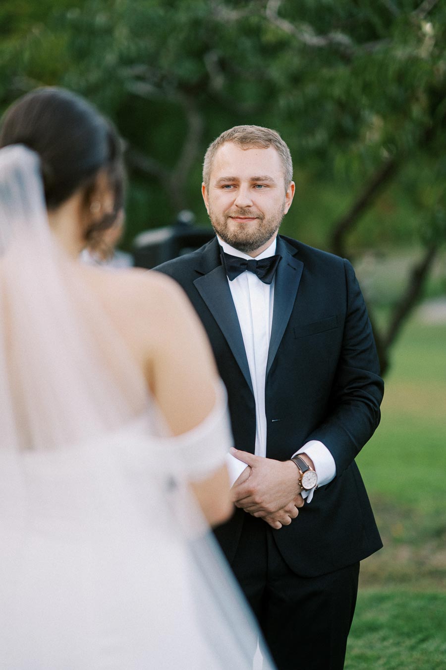 Man in a tuxedo smiles at a bride during an outdoor wedding ceremony, capturing a heartfelt moment with greenery in the background.