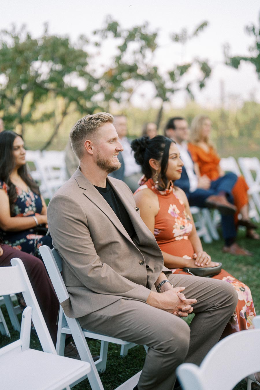 Guests seated outdoors at a wedding ceremony, focusing on a man in a beige suit and a woman in a floral dress sitting attentively in white chairs.