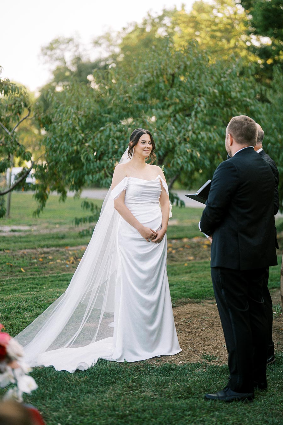 Bride in elegant white dress with veil stands in garden during outdoor wedding ceremony, facing the officiant in a suit.