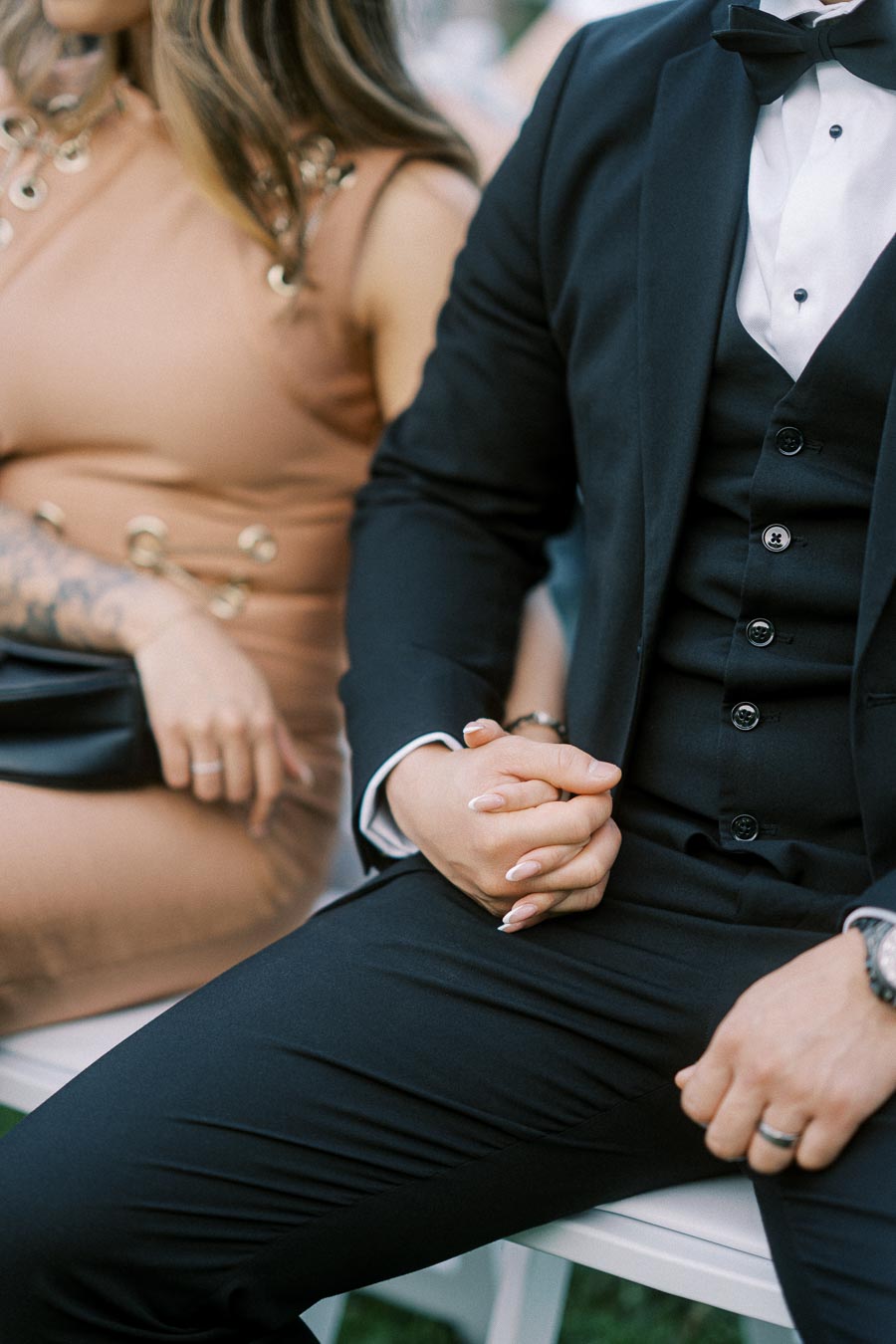 A couple holding hands at an outdoor event, dressed in formal attire with a man wearing a black suit and a woman in a beige dress.
