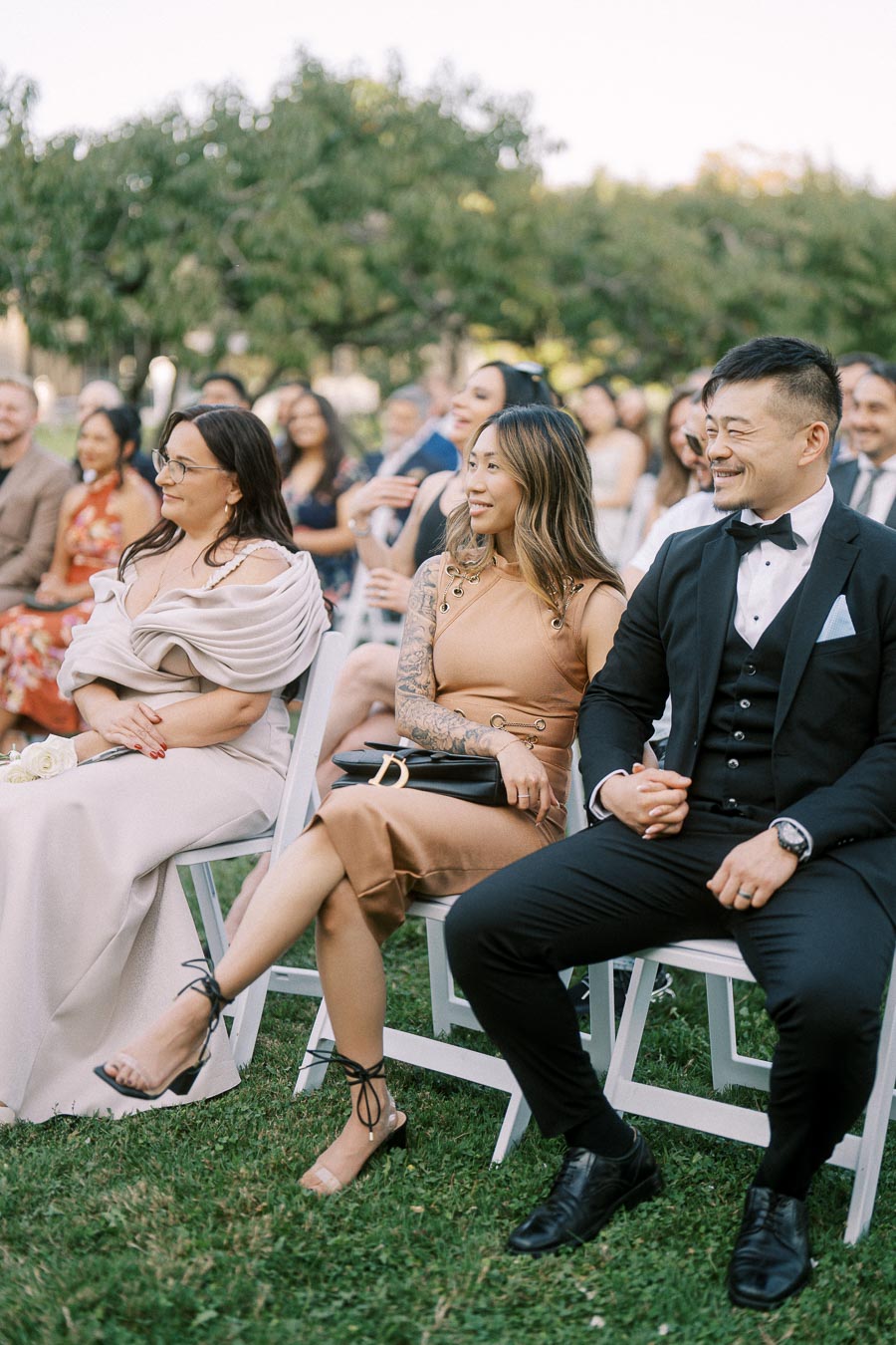 Guests seated outdoors at a formal event, dressed elegantly, enjoying a sunny day. The background features a lush, green garden setting, contributing to the joyful atmosphere.