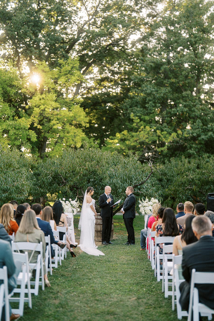 Outdoor wedding ceremony in a lush garden setting, featuring a couple standing before an officiant surrounded by guests seated on white chairs, with sunlight filtering through the trees.