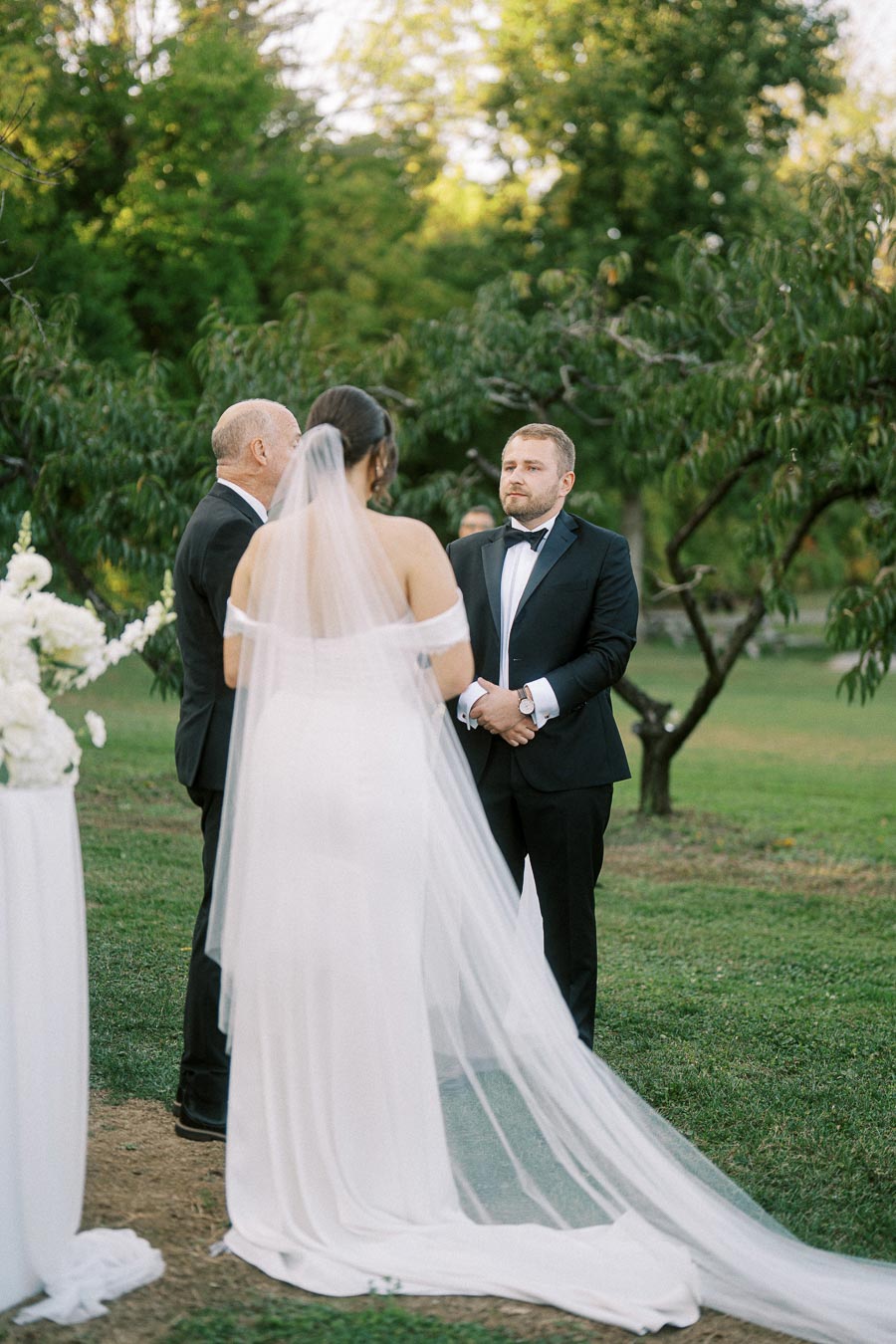 Outdoor wedding ceremony with bride in a white gown and veil standing alongside groom in a suit, surrounded by greenery and floral arrangements.