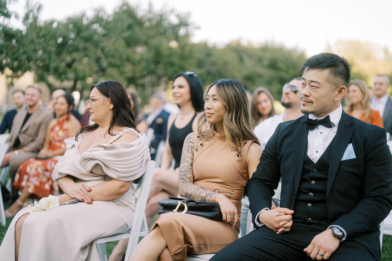 Group of elegantly dressed guests seated at an outdoor wedding ceremony, surrounded by greenery and soft sunlight.
