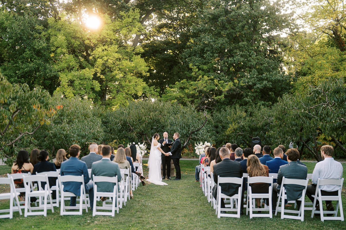 Outdoor wedding ceremony in a lush garden setting, with a couple exchanging vows under a tree as guests watch from white chairs.